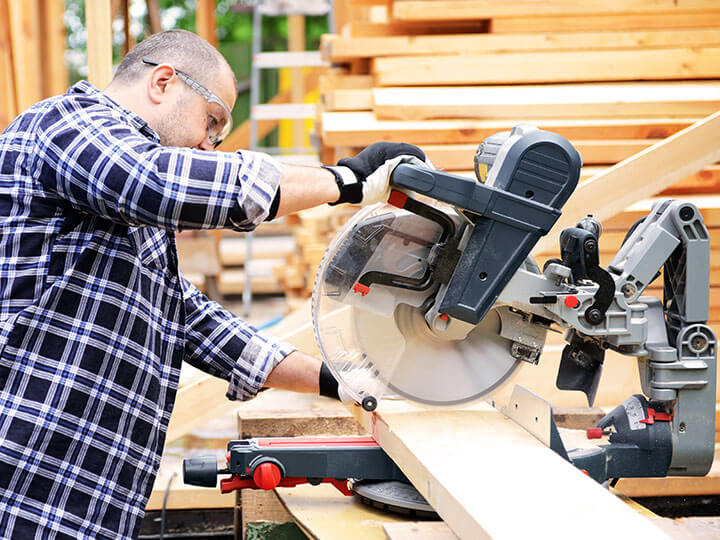 Construction worker in a plaid shirt wearing safety goggles cutting a wooden board with a miter saw